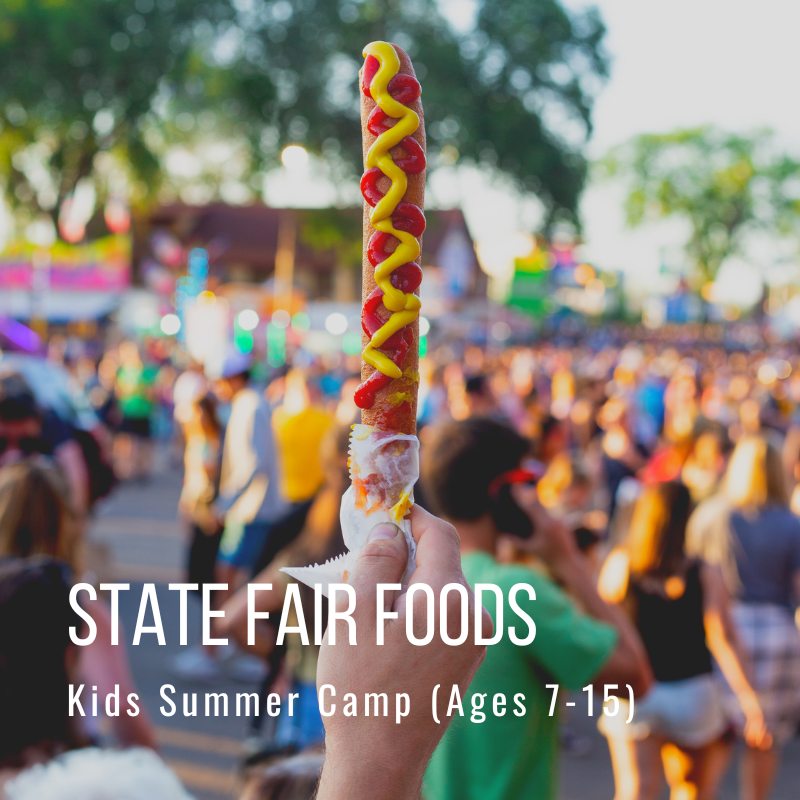 Hand holding corn dog with ketchup and mustard at a fair with background crowd.