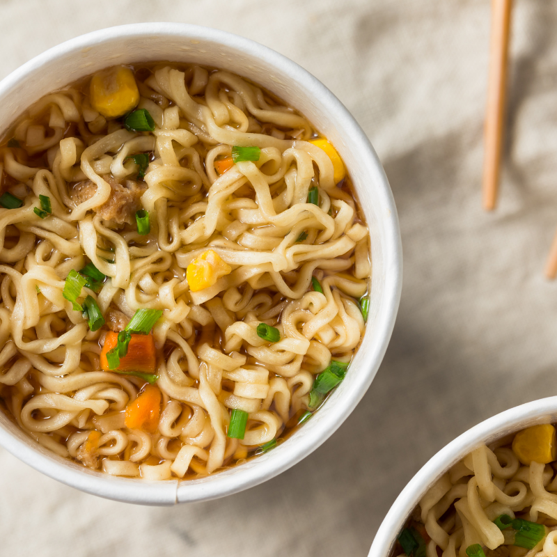 Cup of instant noodles with corn and green onions on a light surface.