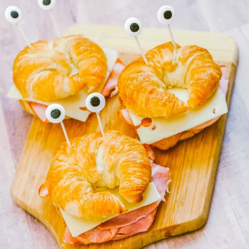Croissant sandwiches with cheese and ham, topped with stick eyes on a wooden board.