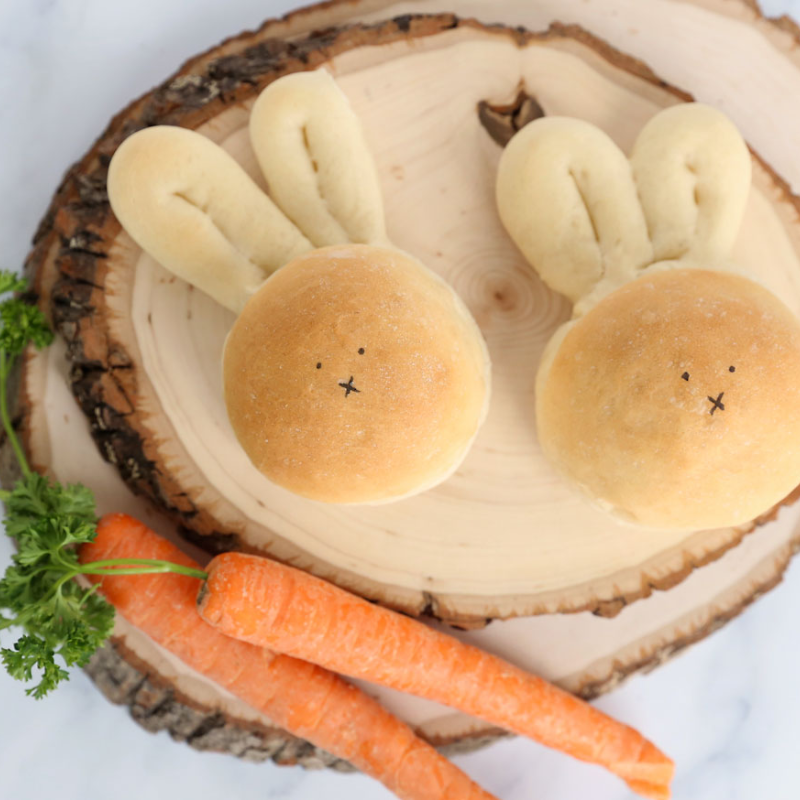 Two bunny-shaped bread rolls with carrots and parsley on wooden slices.