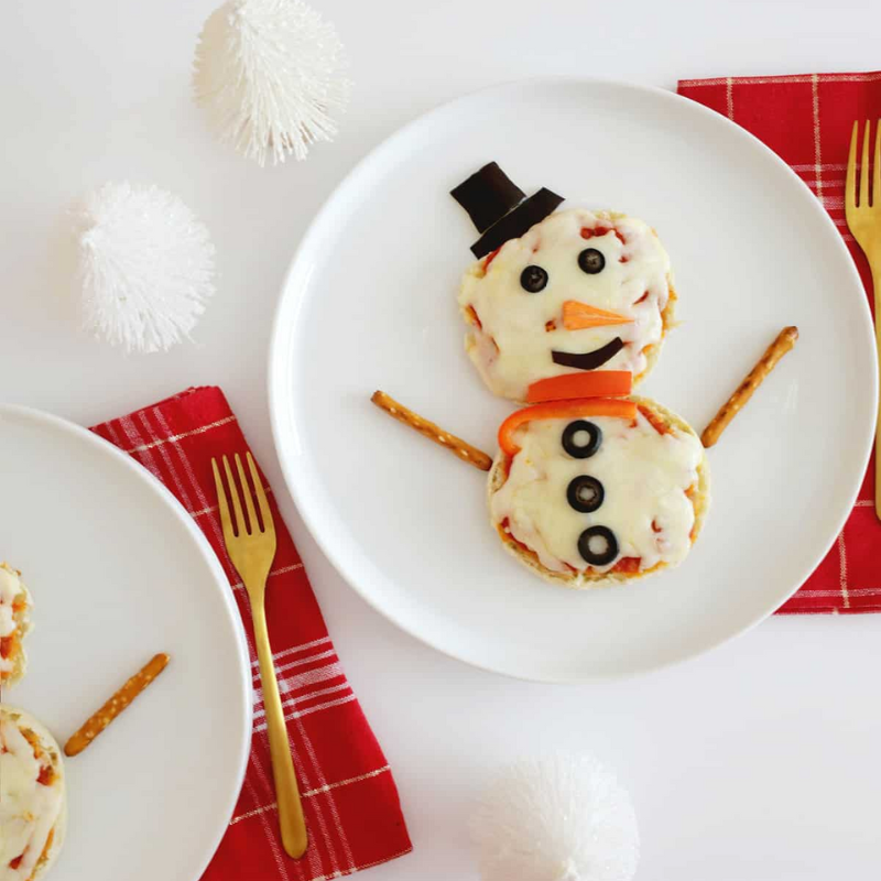 Snowman-shaped pizza with pretzel stick arms on a plate, gold fork, red napkin, white decorations.