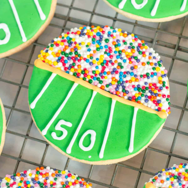 A football-shaped cookie with green icing and sprinkles on one half, resembling a field with a '50' yard line.
