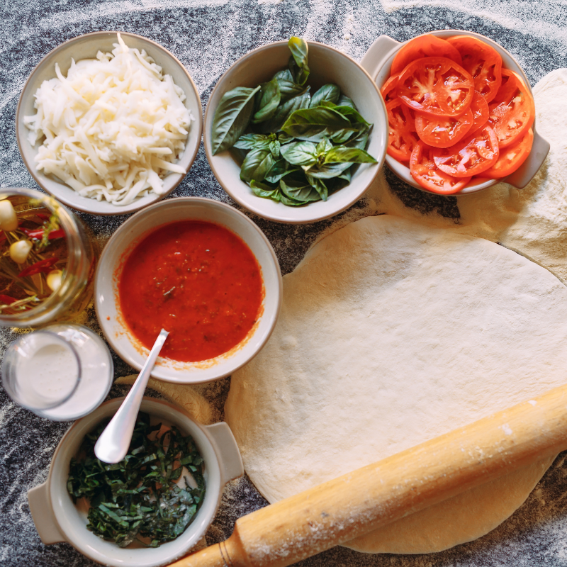 Pizza ingredients with rolled dough, tomato sauce, cheese, basil, and tomatoes on a floured surface.