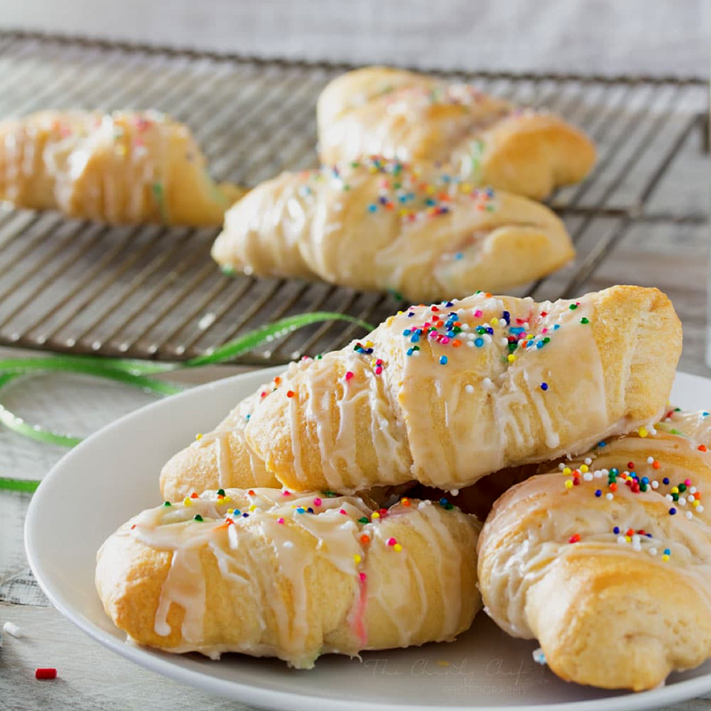 Plate of glazed crescent rolls with sprinkles, cooling rack in background.