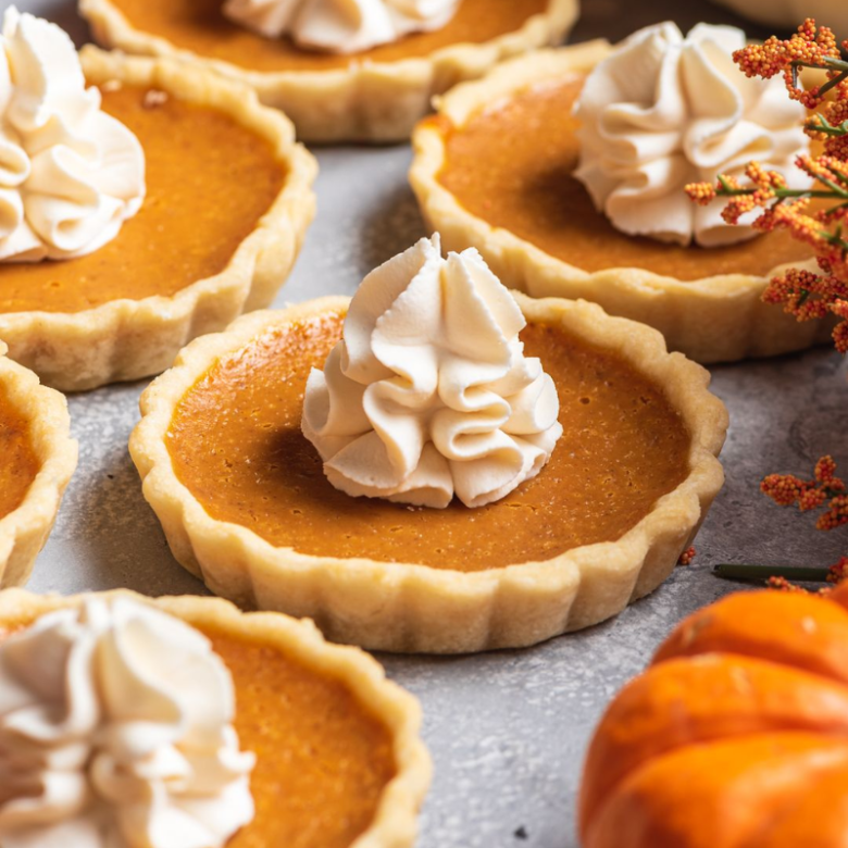 Mini pumpkin pies with whipped cream on top surrounded by autumn decor.