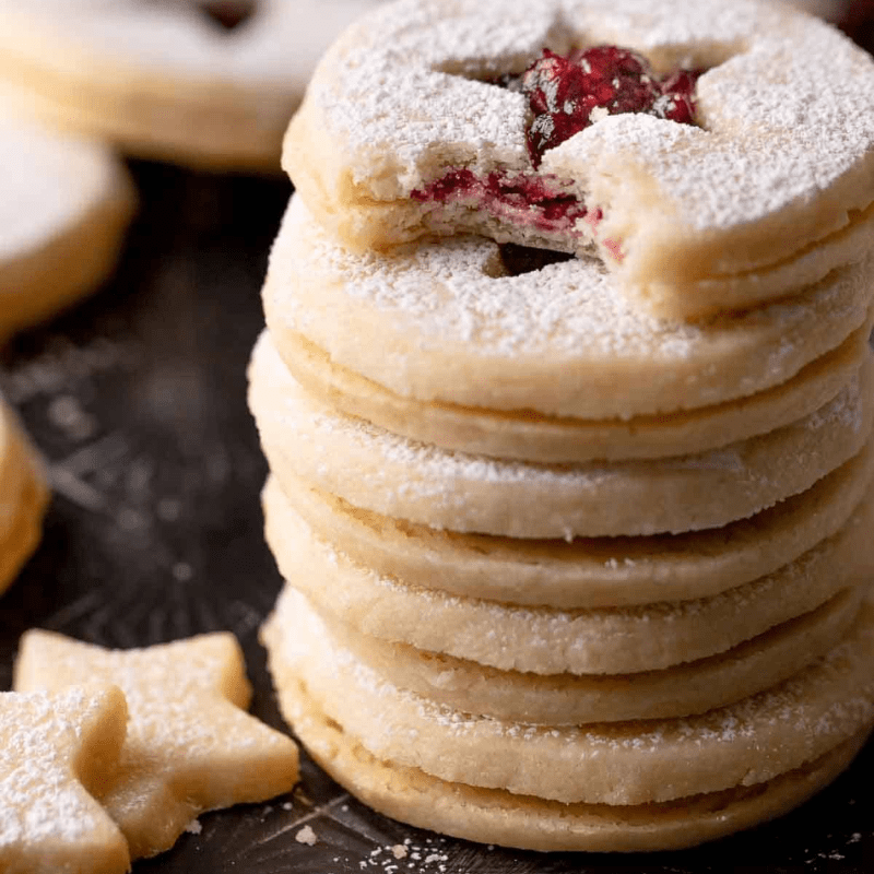 Stack of powdered sugar cookies with berry filling, one with a star-shaped cutout, on a dark background.