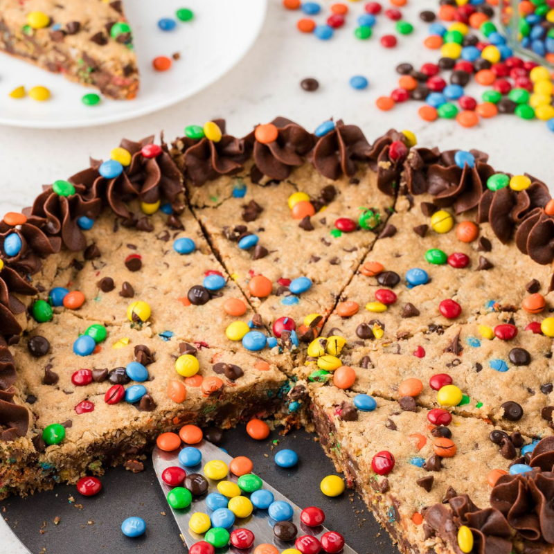 Chocolate chip cookie cake with colorful candies and chocolate frosting, one slice being served.