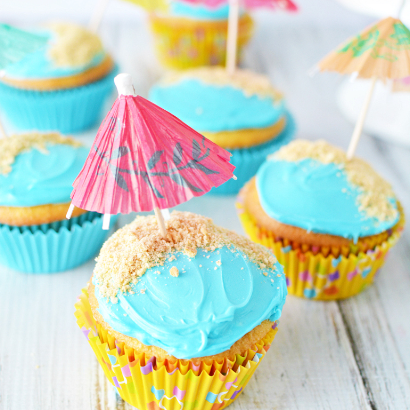 Cupcakes with blue icing, sand sprinkles, and mini umbrellas on top.