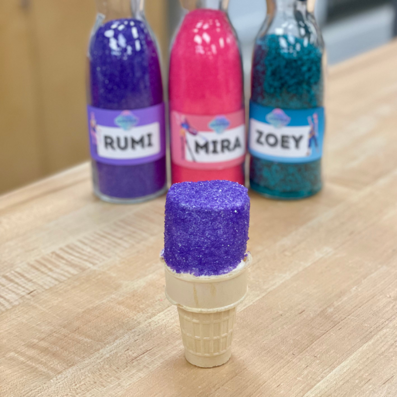 Purple sugar on ice cream cone in focus; bottles labeled Rumi, Mira, Zoey behind.