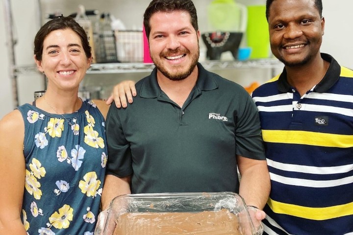 Three people smiling, holding a tray of chocolate dessert in a kitchen setting.