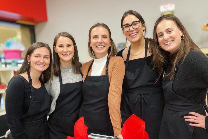 Five women in aprons smiling behind a table with plates of food and red napkins.