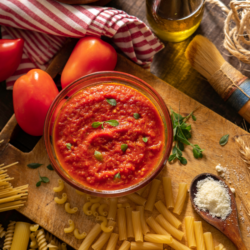Bowl of tomato sauce surrounded by tomatoes, pasta, and herbs on a wooden table.