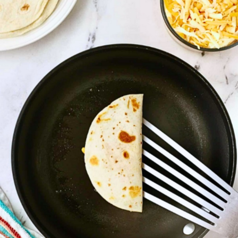 Half-folded tortilla in skillet, spatula underneath, with cheese and tortillas nearby.