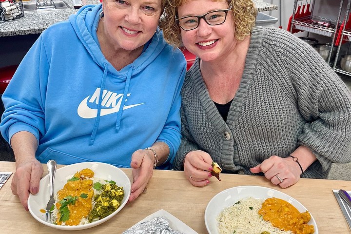 Two women smiling at a table with Indian curry dishes and foil-wrapped food.