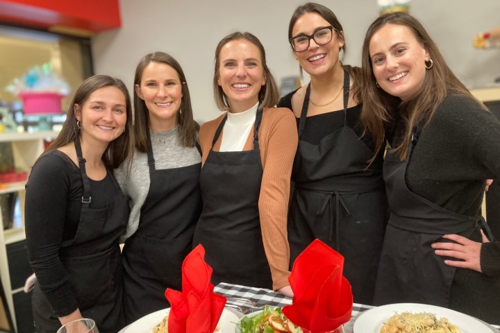 Five women in black aprons smiling at a dining table with red napkins and plates of food.