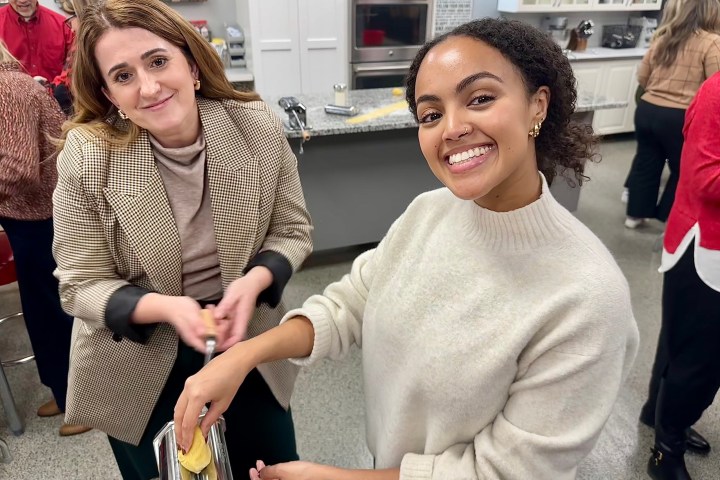 Two women smiling while using a pasta machine in a kitchen setting.