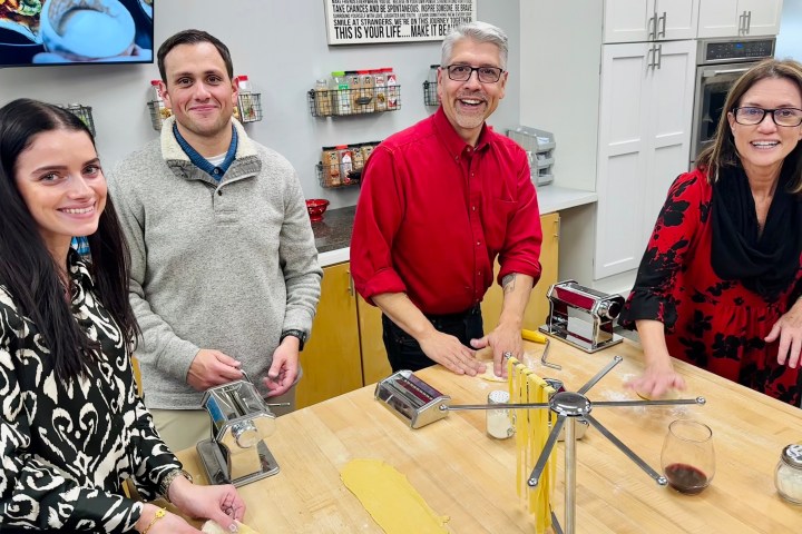 Four people making pasta together in a kitchen, smiling at the camera.