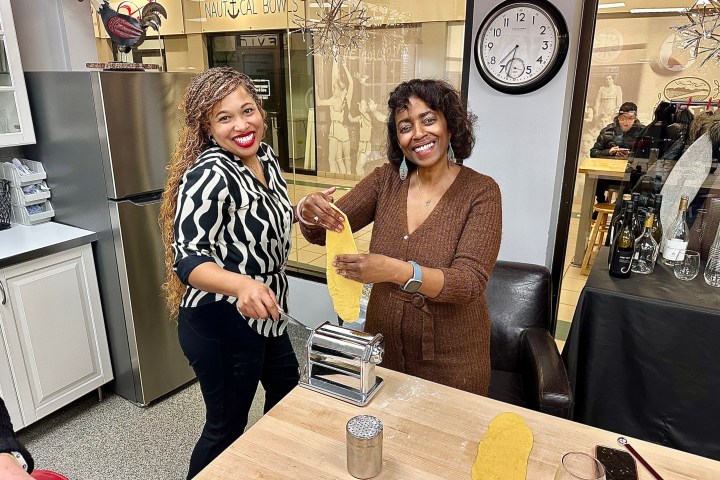 Two women smiling while making pasta with a pasta machine in a kitchen.
