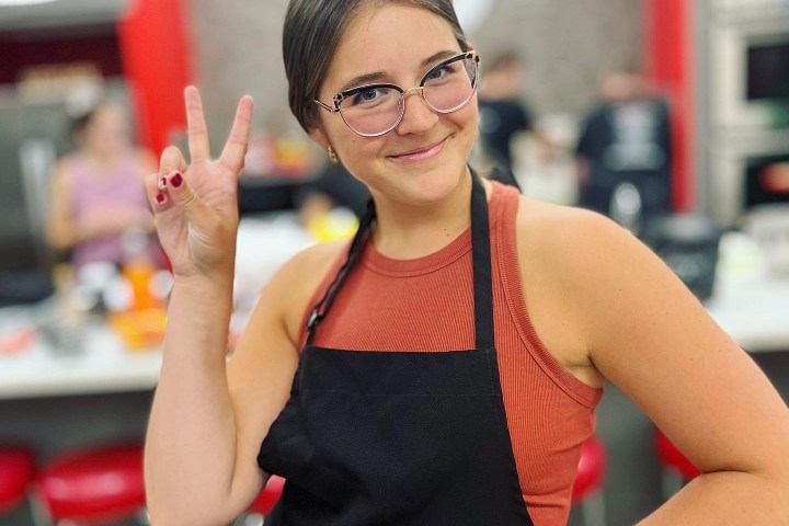 Smiling person in apron, making a peace sign in a kitchen setting.