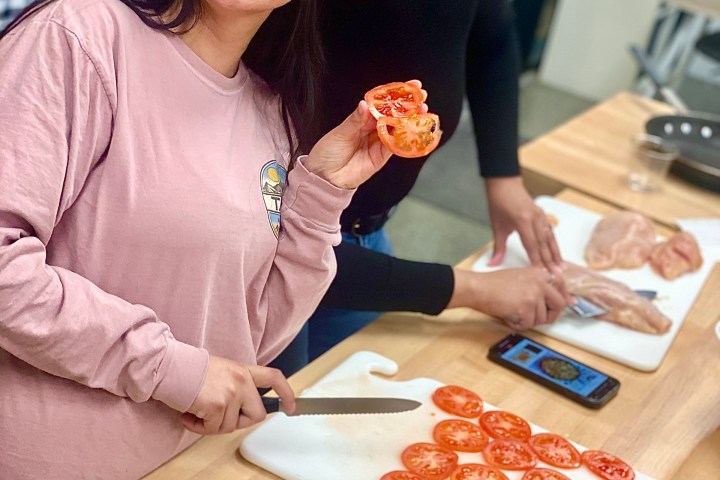Two women smiling while slicing tomatoes on a wooden kitchen counter.