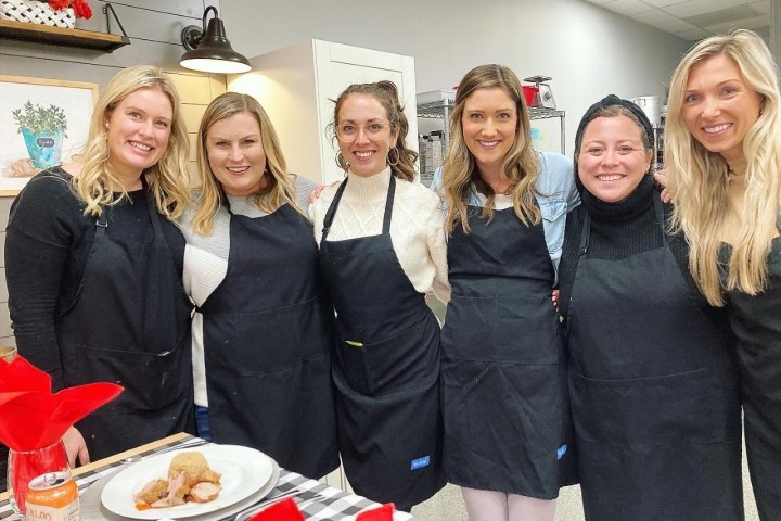 Six women wearing aprons smiling in a kitchen with table set for a meal.