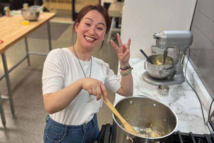 Person smiling and cooking with a pot on a stove, holding a wooden spoon, in a kitchen setting.