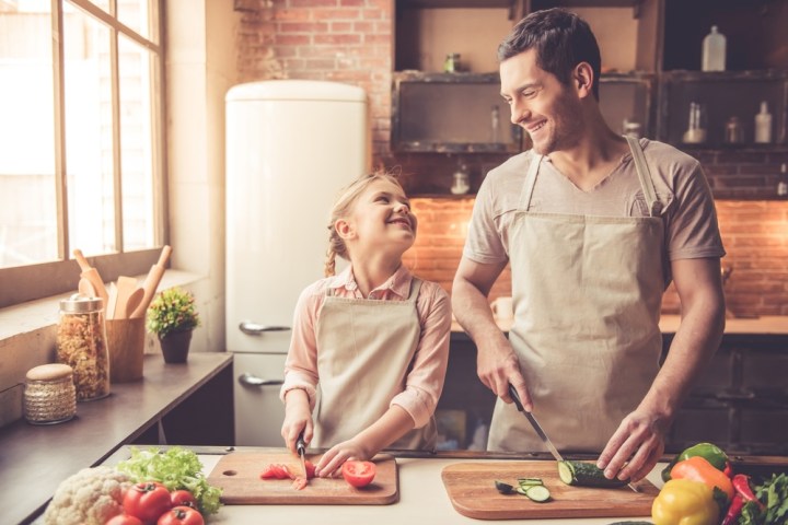Father and daughter smiling while chopping vegetables together in a kitchen.