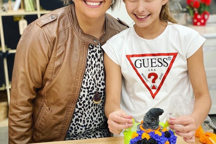 Two people smiling with a colorful cake; one wears a leather jacket, the other a 'Guess' t-shirt.