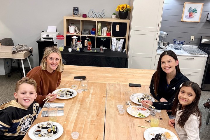 Four people smiling, seated at a table with sushi plates in a kitchen setting.