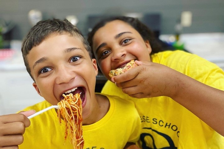 Two kids in yellow shirts enjoying spaghetti and pizza at a table.