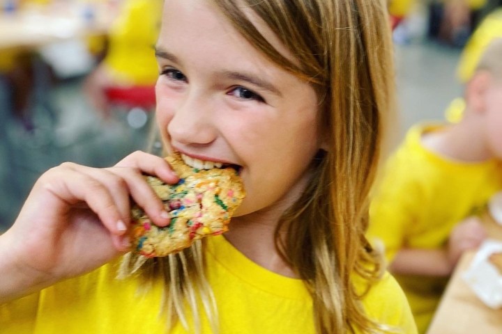 Child in a yellow shirt eating a colorful cookie in a cafeteria.