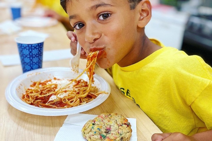 Child in yellow shirt eating spaghetti at a table, with a cookie and paper cup nearby.
