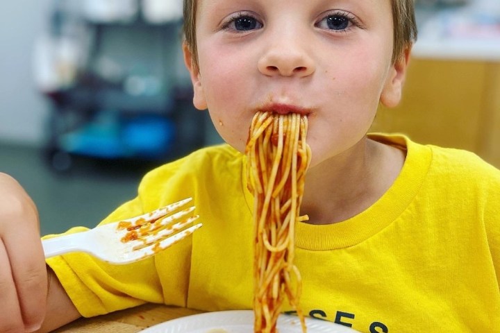 Child eating spaghetti with plastic fork, wearing a yellow shirt.