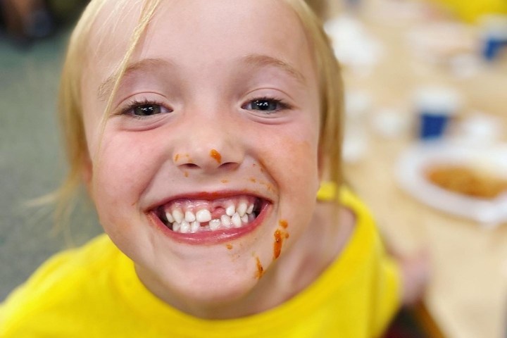 Smiling child with tomato sauce on face, wearing a yellow shirt.