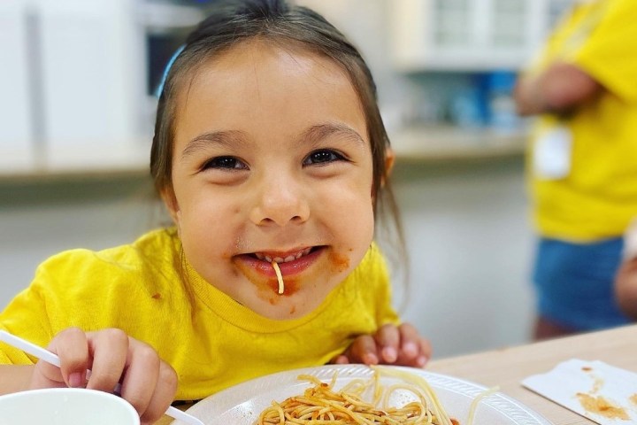 Child smiling with spaghetti sauce on face, holding fork next to plate of spaghetti.