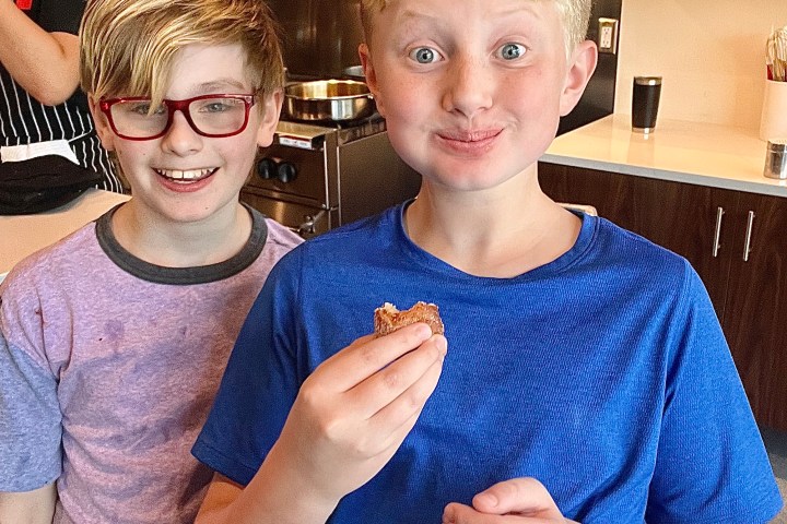 Two smiling boys in a kitchen; one holds a cookie, surrounded by shelves and kitchenware.