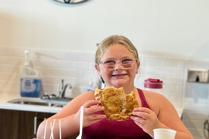 Child holding a sandwich, smiling at a kitchen table with a clock on the wall.