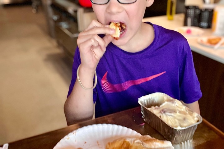 Child with glasses eating pastry in kitchen, wearing purple shirt.