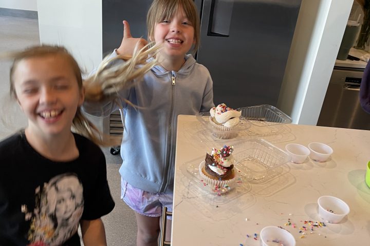 Two children smiling in a kitchen by a counter with cupcakes and sprinkles.