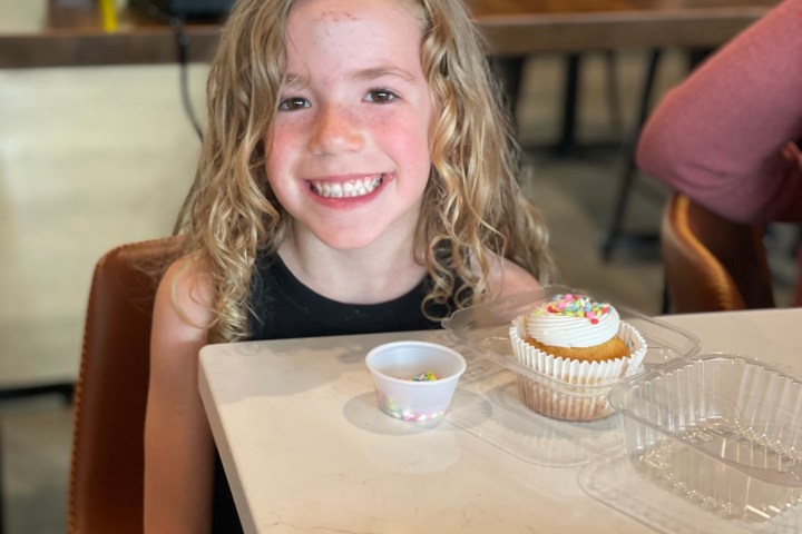Child with curly hair smiling, decorating a cupcake with colorful sprinkles at a table.