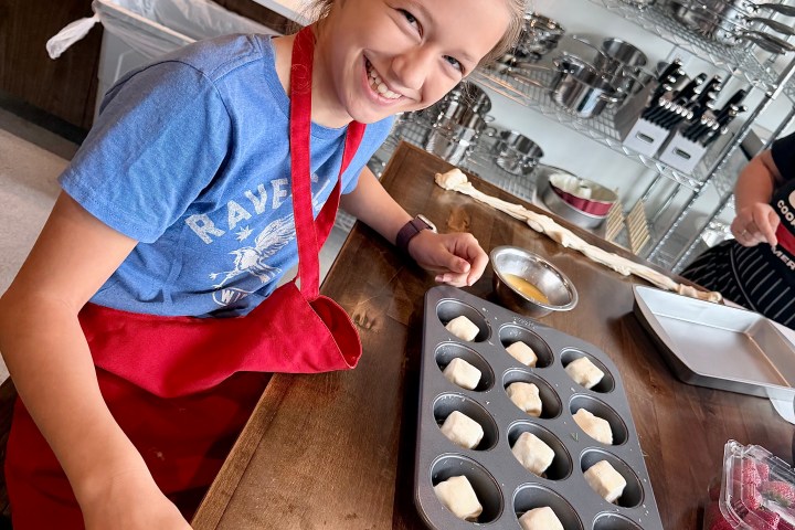 Smiling child in red apron beside a muffin tray on a kitchen counter.