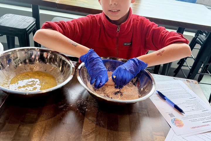 Child in red jacket, wearing blue gloves, mixes ingredients in bowls on a table.