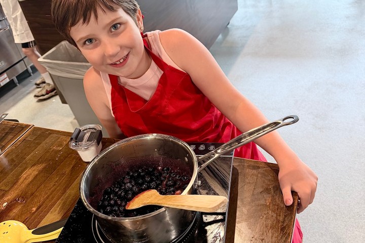 Child in red apron cooking berries in a pot on a kitchen counter.