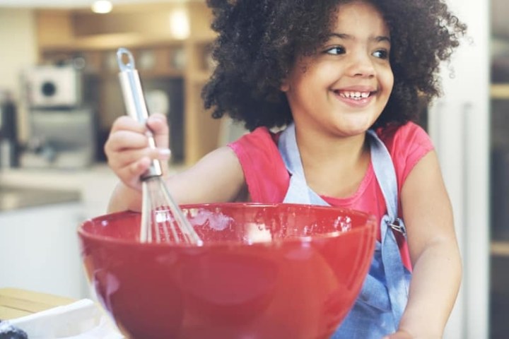 Smiling child with curly hair whisking in a red bowl in a kitchen.