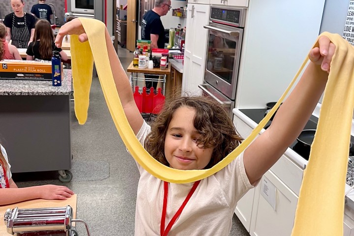 Child in a kitchen holding a sheet of pasta with a pasta machine on the table.