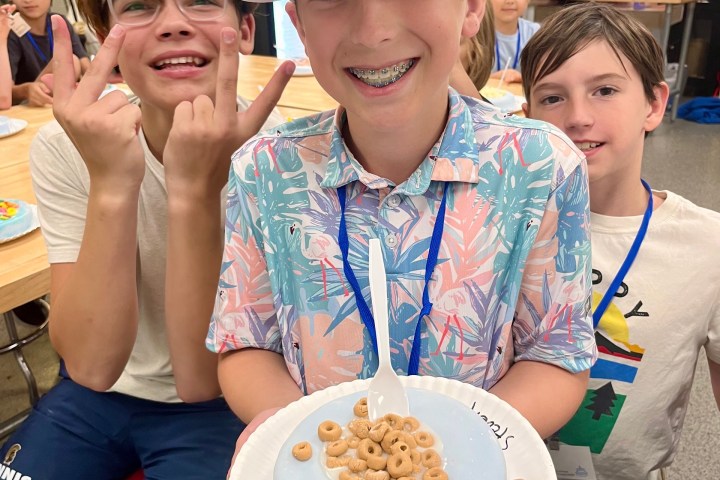 Three kids smiling; one holds a plate with a small cake topped with cereal. Others make peace signs.