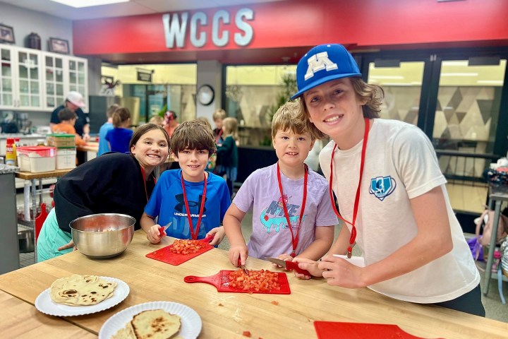 Smiling kids in a kitchen preparing food on a wooden table with cutting boards.