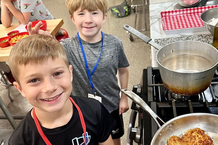 Two boys smiling by a stove with food cooking in a pan.