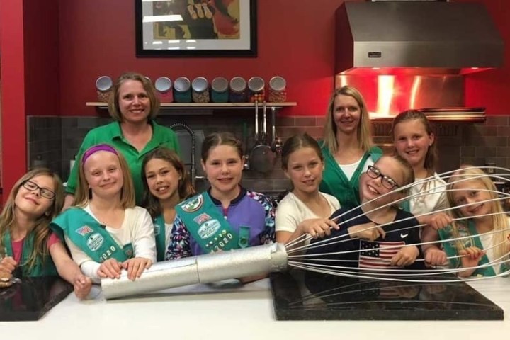 Group of kids and two adults in a kitchen holding a giant whisk.