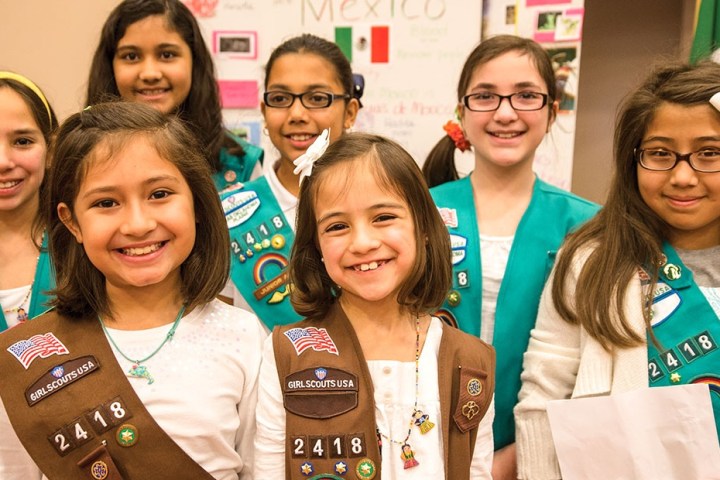Smiling girl scouts in uniforms with badges, standing indoors at an event.
